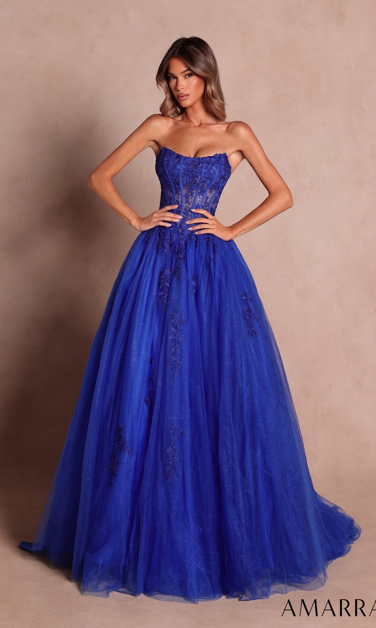 A woman wearing the Amarra 88913 strapless long prom ball gown in royal blue, which features a sheer lace corset and elegant floral embroidery, stands against a plain beige background.